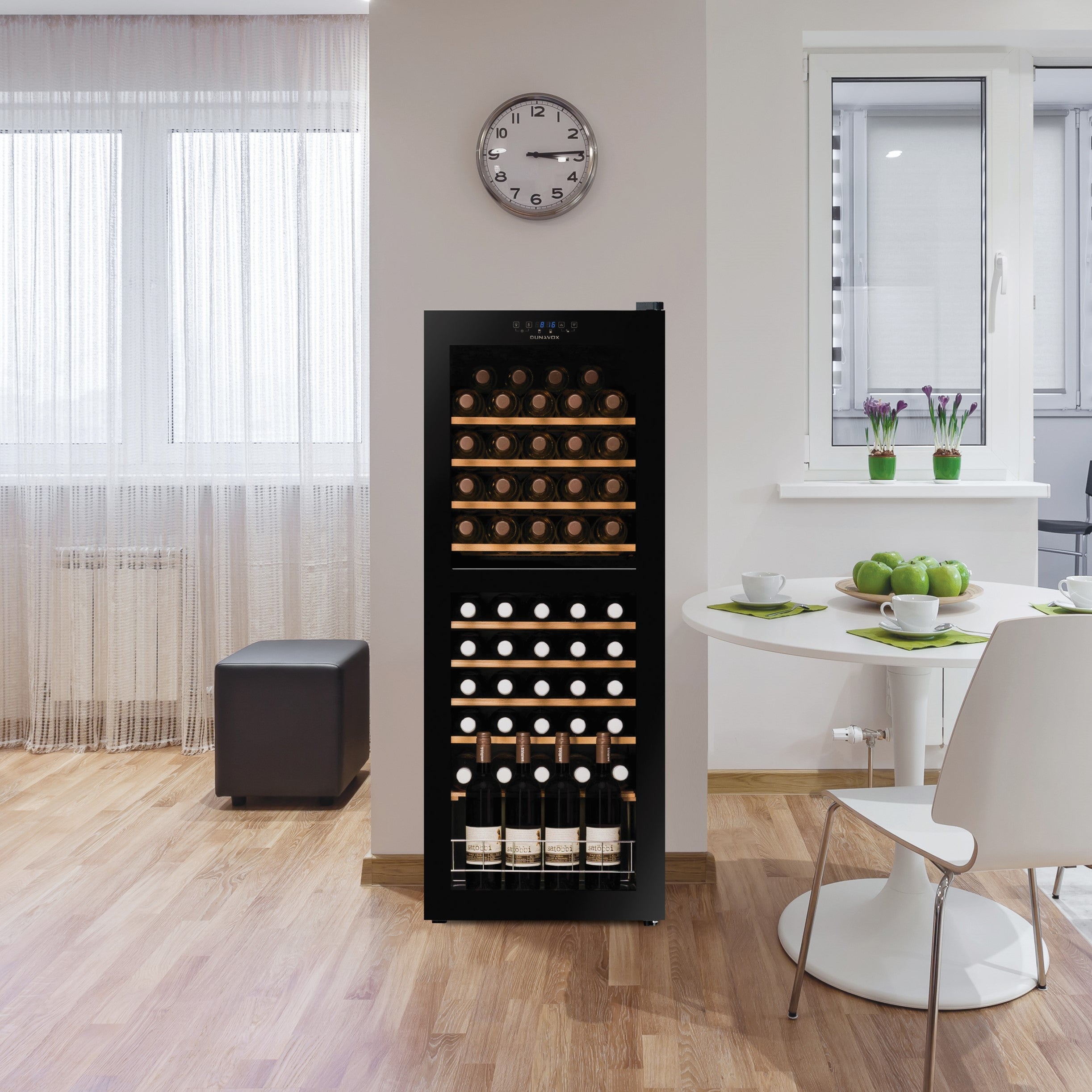 Alt text: Intérieur moderne avec un cellier à vin noir vertical rempli de bouteilles, près d’une table ronde blanche et d’un grand fenêtre, décor minimaliste et plantes vertes sur le rebord. montre murale au-dessus. Intègre le mot-clé: quel est le meilleur système de conservation du vin en europe.