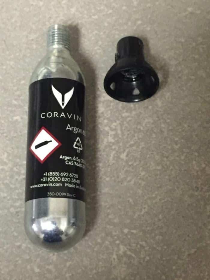 Bottiglia di gas enologico Ar-gon Coravin con etichetta nera, tappo copricapsula staccato accanto, contenitore trasparente su piano grigio. Quale gas enologico usare per conservare il vino aperto.