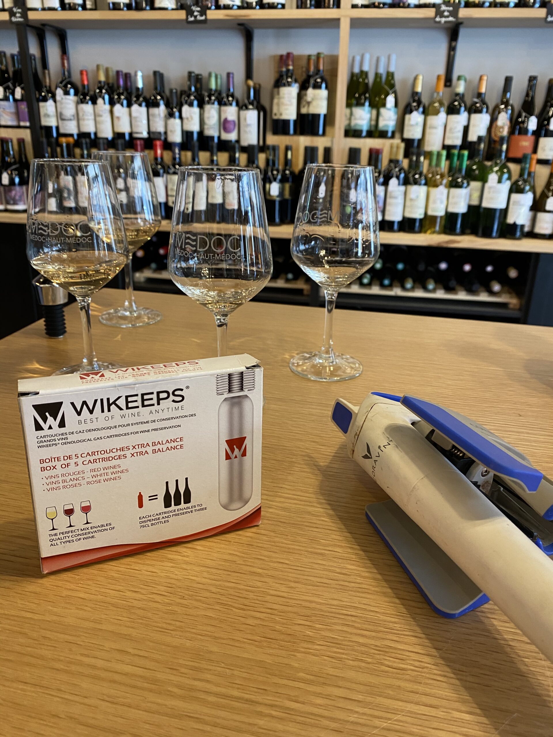 Three wine glasses on a wooden bar with assorted bottles in the background; a boxed WIKEEPS wine preservation kit and a handheld device rest on the counter.